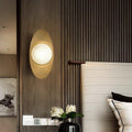A modern bedroom with chic elegance features a glowing Metal Boat-Shaped Wall Light and ribbed gray paneling, complemented by a white upholstered headboard. A black vase with white flowers sits on the bedside table, while a black-and-white portrait and contemporary lighting complete the sophisticated look.