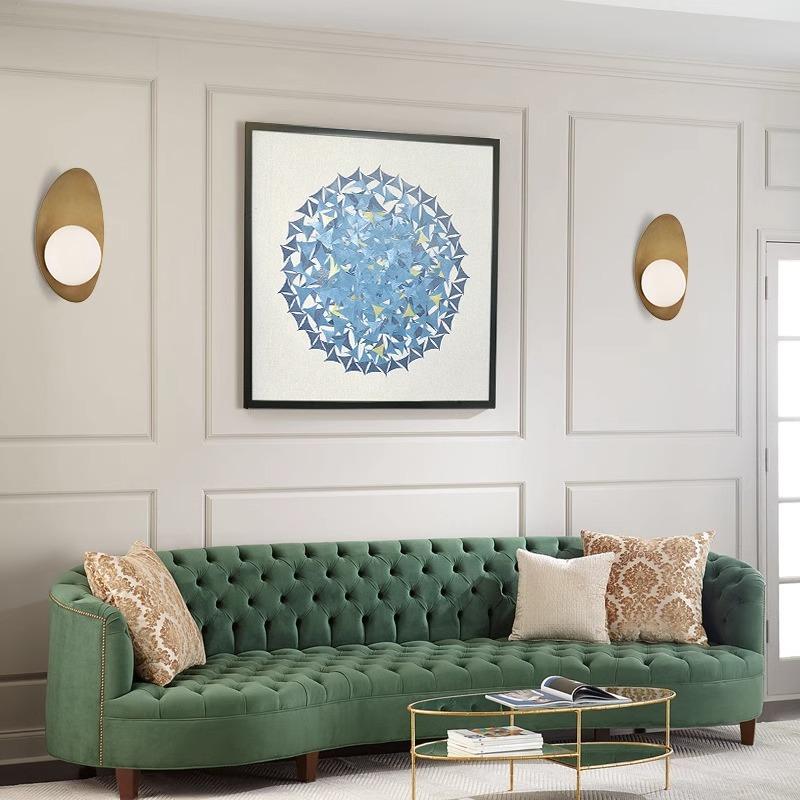 A chic living room showcases a green velvet tufted sofa adorned with patterned pillows, geometric blue artwork on the wall, and modern Metal Boat-Shaped Wall Lights. A glass coffee table rests on a light floor.