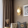 A modern living room exudes chic elegance with a beige sofa, an embroidered pillow, and a metal boat-shaped wall light mounted next to abstract art. A small round table holds a dark vase with dried flowers, while floor-length brown curtains add warmth nearby.