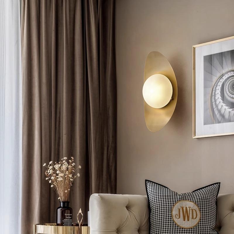 A modern living room exudes chic elegance with a beige sofa, an embroidered pillow, and a metal boat-shaped wall light mounted next to abstract art. A small round table holds a dark vase with dried flowers, while floor-length brown curtains add warmth nearby.