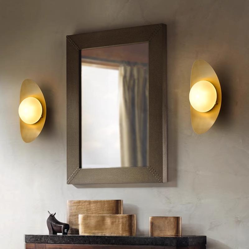A rectangular mirror with a dark frame hangs on a light wall, flanked by Metal Boat-Shaped Wall Lights. Below, a dark shelf holds small decor items like a sculpture and books, enhancing the spaces modern-style lighting and chic elegance.