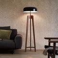 A modern Tripod Floor Lamp with a black shade and wooden legs stands next to a dark gray sofa adorned with a green cushion. The room features textured beige walls, a wooden floor, and a partially visible wooden chair.