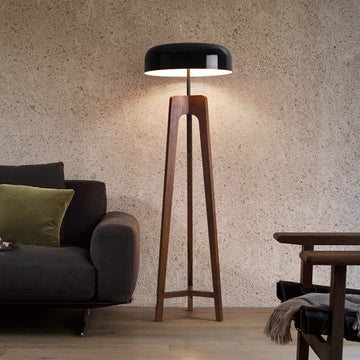 A modern Tripod Floor Lamp with a black shade and wooden legs stands next to a dark gray sofa adorned with a green cushion. The room features textured beige walls, a wooden floor, and a partially visible wooden chair.