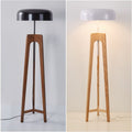 Two Tripod Floor Lamps with wooden bases: the left with a black shade, unlit; the right with a white shade, casting a warm glow. Both feature minimalist designs against a white background.