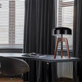 A Tripod Floor Lamp with a black shade and wooden legs stands on a minimalist gray desk. An open book lies nearby. The room features dark curtains and light-filtering blinds on the windows, with a black chair close by.