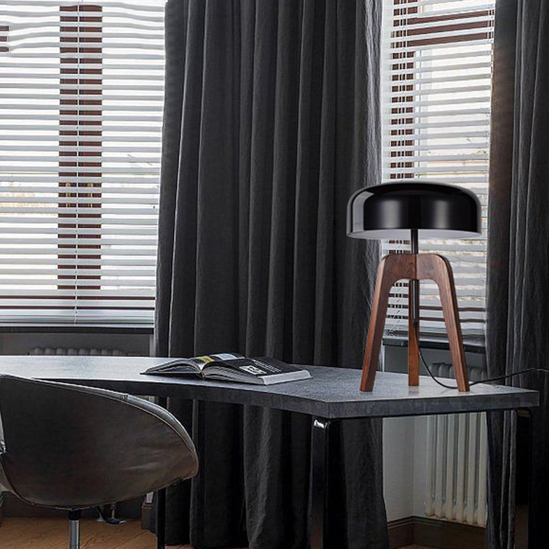 A Tripod Floor Lamp with a black shade and wooden legs stands on a minimalist gray desk. An open book lies nearby. The room features dark curtains and light-filtering blinds on the windows, with a black chair close by.