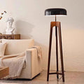 A modern living room showcases the Tripod Floor Lamp, featuring a wooden frame and black dome-shaped shade. It stands near a white couch adorned with a light-colored blanket, with a decorative bust placed on the wooden cabinet behind.