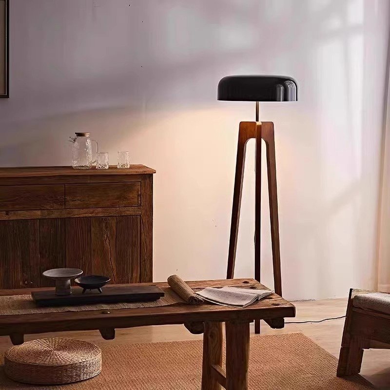 A cozy room features a wooden table, bench, and cabinet. The Tripod Floor Lamp with a black shade casts a warm glow. A woven mat and glass pitchers enhance the rustic decor against a softly lit wall.
