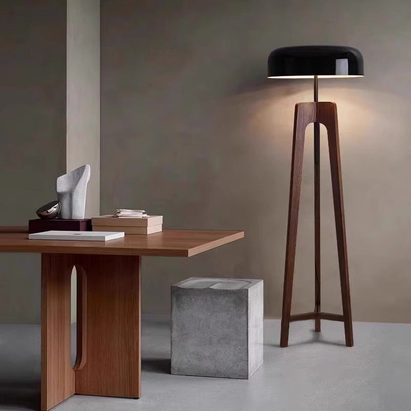 A modern interior includes a wooden table with a sculpture and books, paired with the tall Tripod Floor Lamp featuring a black lampshade. A square concrete stool sits beside the table, with the wall painted in neutral tones.