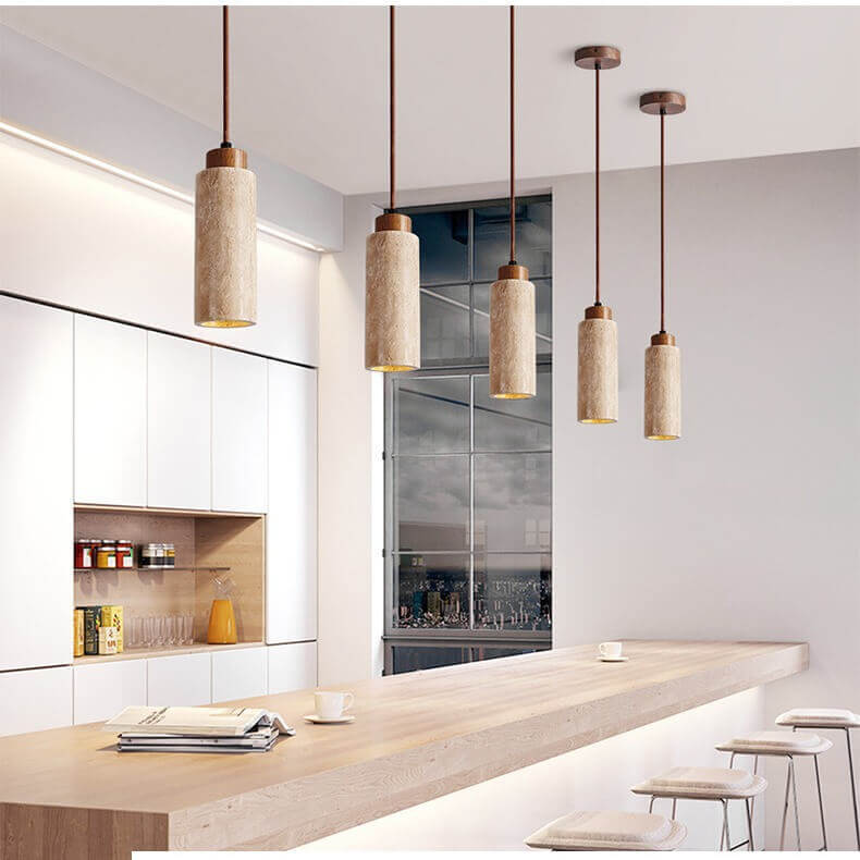 A minimalist dining area showcases a round table blending travertine and wood with beige walls. Above, three Travertine Restaurant Chandeliers provide dimmable LED lighting. A white vase of dried branches and a small sculpture adorn the table, flanked symmetrically by two wooden doors.