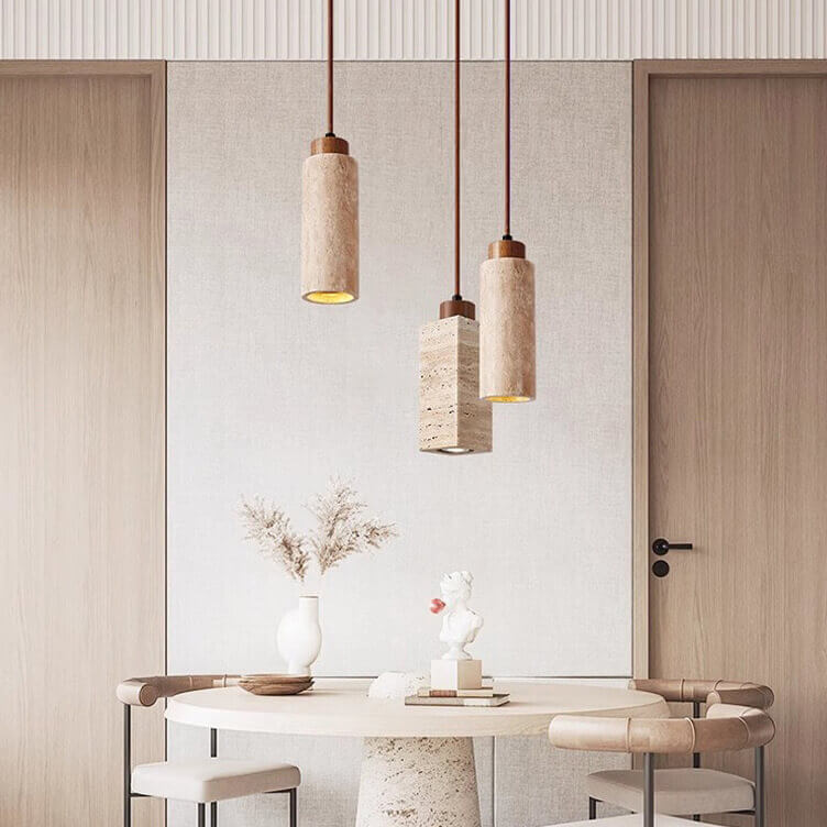 A minimalist dining area showcases a round table blending travertine and wood with beige walls. Above, three Travertine Restaurant Chandeliers provide dimmable LED lighting. A white vase of dried branches and a small sculpture adorn the table, flanked symmetrically by two wooden doors.
