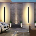 A sleek outdoor space features a luxury car on patterned pavement, while two Outdoor Linear Wall Lights (IP65 waterproof) illuminate the concrete wall, bordered by potted greenery. A cushioned seating area in the foreground completes the sophisticated ambiance.