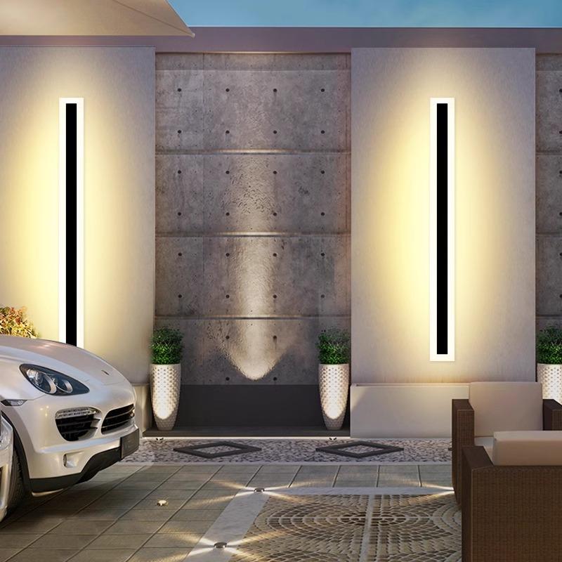 A sleek outdoor space features a luxury car on patterned pavement, while two Outdoor Linear Wall Lights (IP65 waterproof) illuminate the concrete wall, bordered by potted greenery. A cushioned seating area in the foreground completes the sophisticated ambiance.
