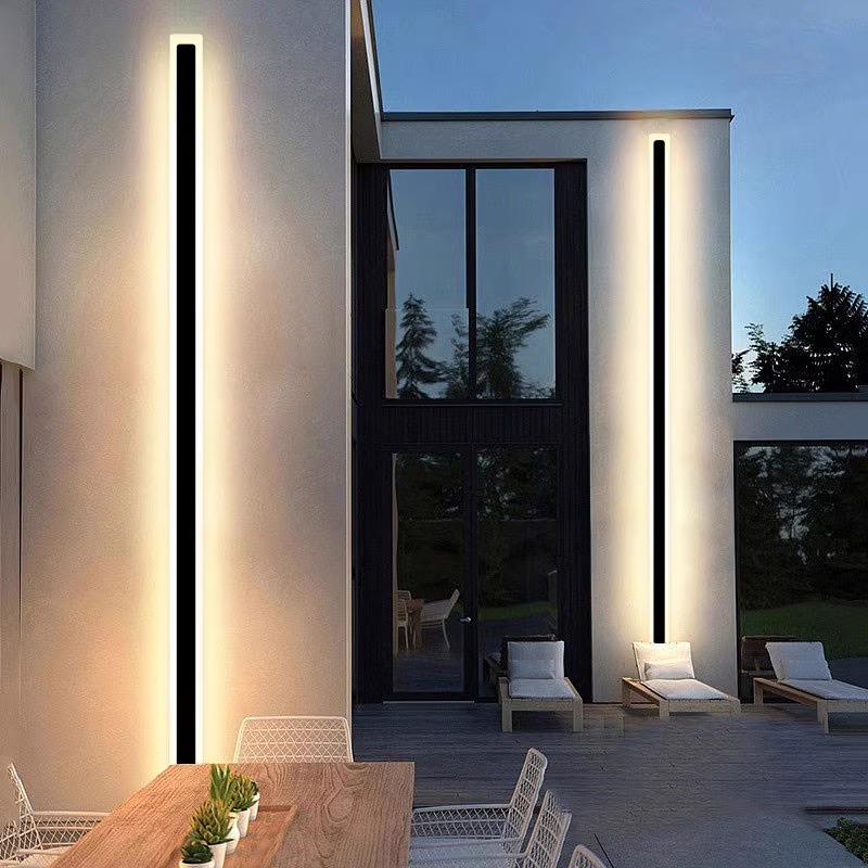 A modern outdoor patio features sleek Outdoor Linear Wall Lights on a two-story building. A wooden dining table with white mesh chairs sits in the foreground, while glass doors and windows reflect trees, and a grassy area can be seen in the background. The lights are IP65 waterproof.