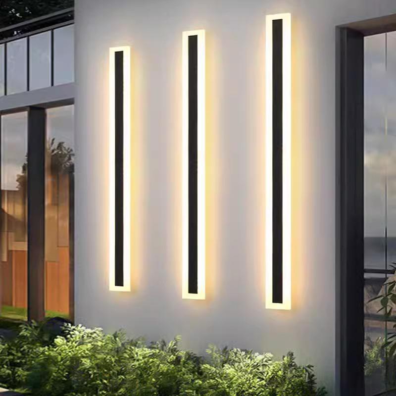 Three Outdoor Linear Wall Lights are evenly spaced on the buildings facade, emitting a warm glow that illuminates the greenery. Reflections of trees and a clear sky appear in the nearby glass door, highlighting their sleek, modern, IP65 waterproof elegance.