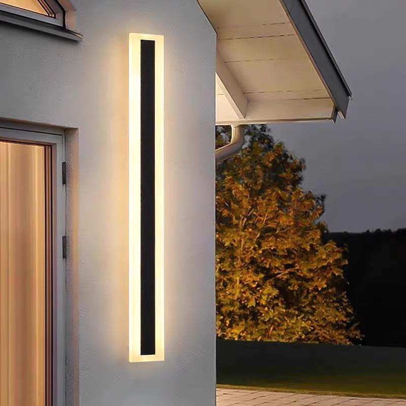 An Outdoor Linear Wall Light with a glowing border is mounted on the house exterior. Its sleek, rectangular design and IP65 waterproof white finish blend harmoniously with the lush trees in the evening backdrop.