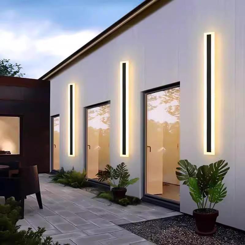 The sleek, modern house exterior showcases three Outdoor Linear Wall Lights with an IP65 waterproof rating. Large glass doors mirror lush greenery, and potted plants line the light gray tiled path, ensuring lasting brilliance against the evening sky backdrop.