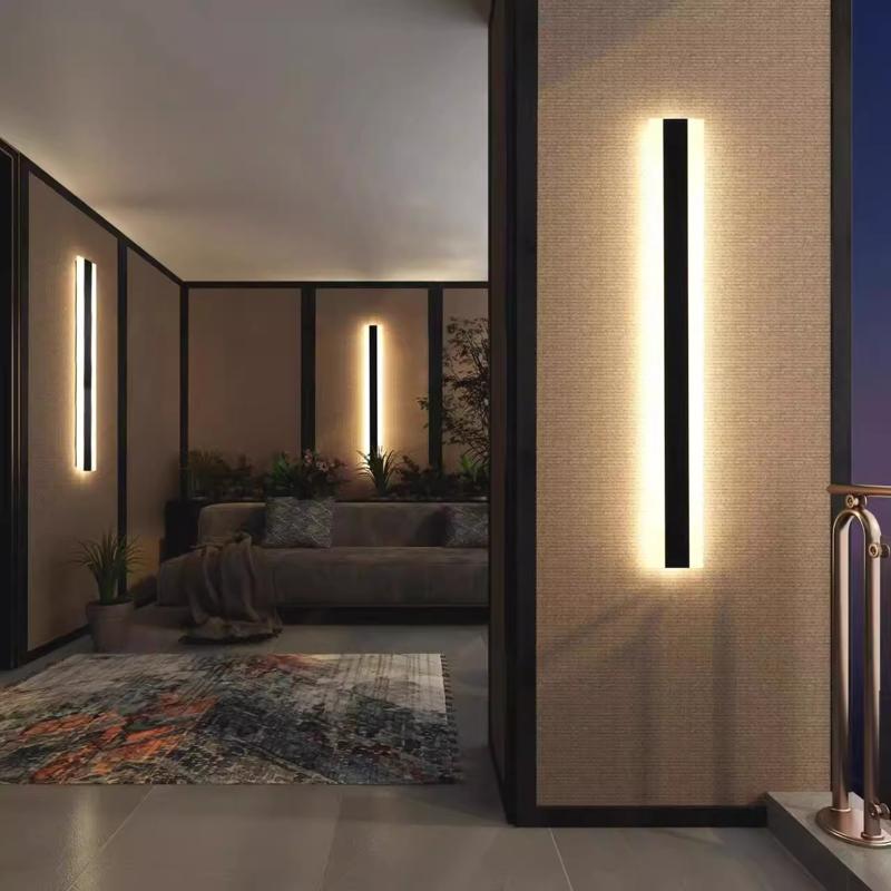 A living room features Outdoor Linear Wall Light with an IP65 rating, casting warmth on textured walls. A gray sofa accented by plants and cushions sits on an abstract rug, while the window offers a breathtaking view of the nighttime sky.