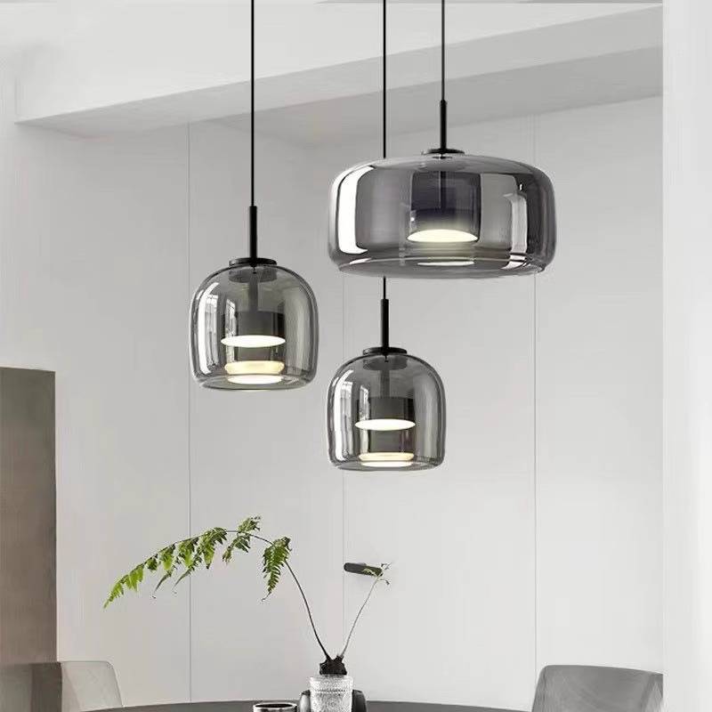Three Retro Chandeliers with translucent glass shades elegantly hang from adjustable wires. Below, a sleek black table with a vase and leafy branch completes the scene. The ambiance exudes simplicity and warmth, accentuated by warm white light against pristine white walls.