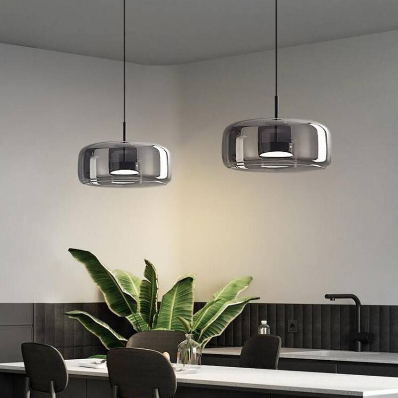 In the modern kitchen, two Retro Chandeliers with glass shades hang above the countertop, their adjustable wires adding versatility. Large green leaves decorate the counter, while black chairs complement the white and dark cabinetry along with a black faucet.