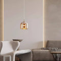 In a modern interior with minimalist style, a sleek gray sofa sits alongside a smooth, curved chair. Light softly fills the space from the Retro Chandelier, featuring a glass shade and adjustable hanging wire. A plaid blanket is casually draped over the sofa for added comfort.