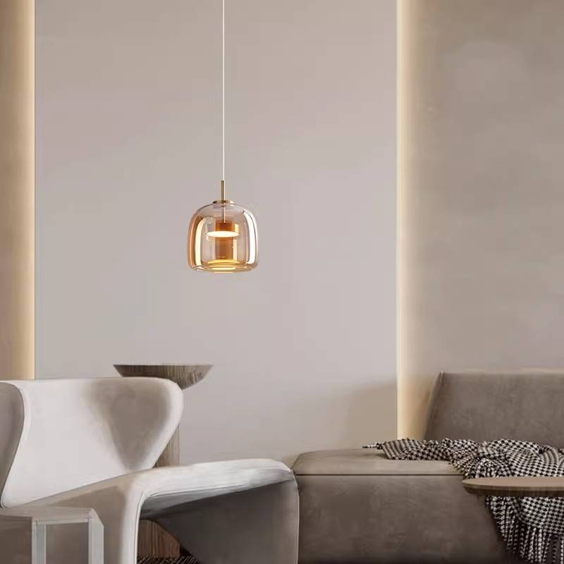 In a modern interior with minimalist style, a sleek gray sofa sits alongside a smooth, curved chair. Light softly fills the space from the Retro Chandelier, featuring a glass shade and adjustable hanging wire. A plaid blanket is casually draped over the sofa for added comfort.
