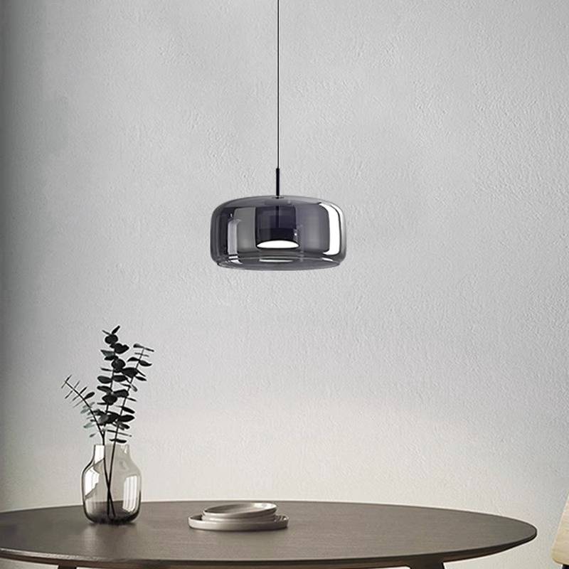 A modern dining scene features the Retro Chandelier, a black glossy pendant lamp with an adjustable wire, over a round wooden table. A glass vase with minimalist branches and a ceramic plate enhance the warm white glow against the light gray wall.