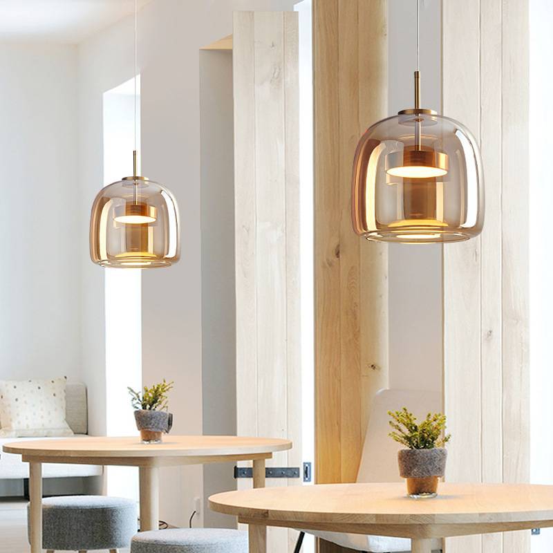 The modern dining area features two round tables, each with a small potted plant. Overhead are Retro Chandeliers with adjustable wires that cast a warm white glow. Wooden panels and minimalist decor in the background create a cozy atmosphere.
