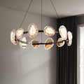The round chandelier, with circular glass-like elements on a metal ring, hangs from the ceiling. It includes smart dimming to adjust to natural light from a nearby window, softly illuminating the room and showcasing its intricate design.
.