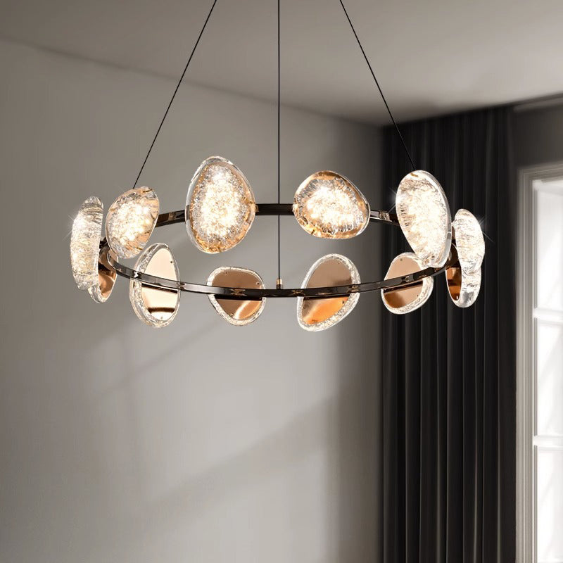 The round chandelier, with circular glass-like elements on a metal ring, hangs from the ceiling. It includes smart dimming to adjust to natural light from a nearby window, softly illuminating the room and showcasing its intricate design.
.
