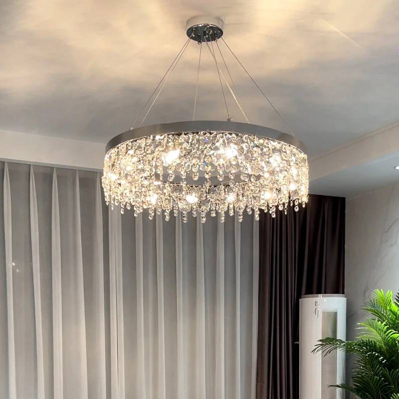 The Crystal Butterfly Chandelier, featuring K9 crystals in a modern circular design, emits soft white light from the ceiling. Its cascading form adds luxury alongside sheer curtains, a tall plant, and a subtle color palette with adjustable lighting perfect for any mood.