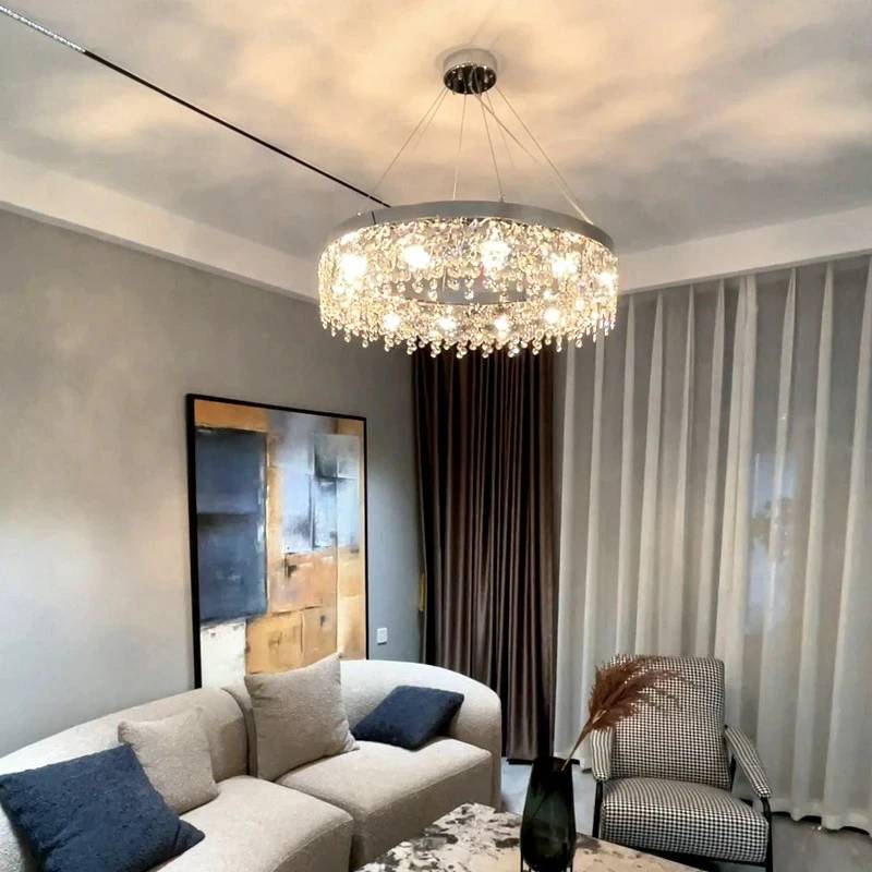 A modern living room showcases a beige sectional sofa with dark cushions, the Crystal Butterfly Chandelier adorned with K9 crystals elegantly hanging from the ceiling. A contemporary abstract painting accents a gray wall, complemented by sheer curtains on the window.