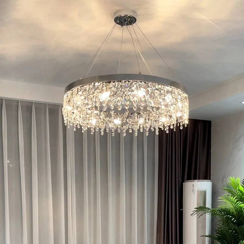 The elegant Crystal Butterfly Chandelier with cascading K9 crystals hangs from the ceiling, casting illuminated reflections on the walls of a room adorned with light-colored curtains, pastel walls, and a corner potted plant.
