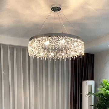 The elegant Crystal Butterfly Chandelier with cascading K9 crystals hangs from the ceiling, casting illuminated reflections on the walls of a room adorned with light-colored curtains, pastel walls, and a corner potted plant.
