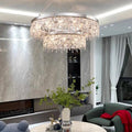 A stylish living room is accentuated with a Crystal Butterfly Chandelier made of K9 crystals, offering adjustable lighting. It complements the marble accent wall, a cozy green armchair, a round table adorned with green foliage, and sheer white curtains by the large window.