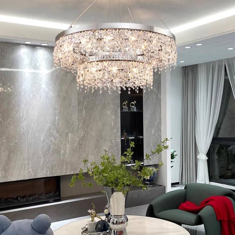 A stylish living room is accentuated with a Crystal Butterfly Chandelier made of K9 crystals, offering adjustable lighting. It complements the marble accent wall, a cozy green armchair, a round table adorned with green foliage, and sheer white curtains by the large window.