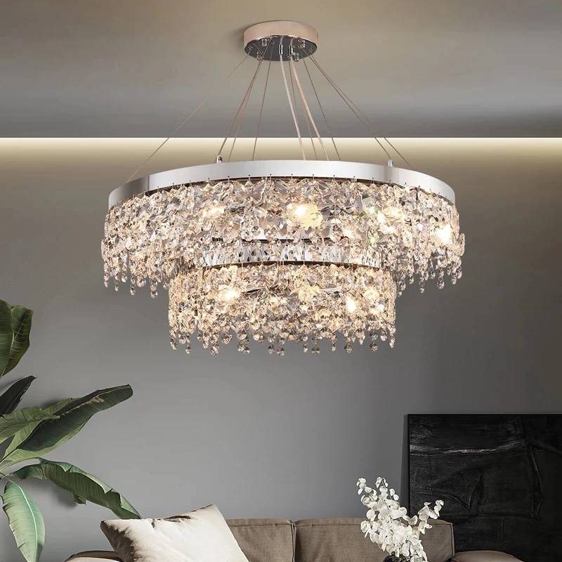 The Crystal Butterfly Chandelier, adorned with cascading K9 crystals, casts a warm glow above a modern grey couch. White cushions and green plants enhance the elegance of the space, while its adjustable lighting adds both style and function.