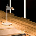 A Simple Desktop Lamp casts light on papers on a wood-grain table. Vertical white light strips in the background against a dark surface emphasize the minimalist design and create a chic ambiance.