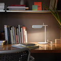 A tidy wooden desk showcases a Simple Desktop Lamp, an open notebook, and a glass of water. Shelves above hold various books, with a potted plant partially visible on the right. Warm LED lighting creates a cozy minimalist ambiance.