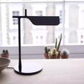 The Simple Desktop Lamp, boasting a sleek black design with LED lighting, sits by a large window lined with small potted cacti. A blurred cityscape forms the backdrop, capturing minimalist elegance.