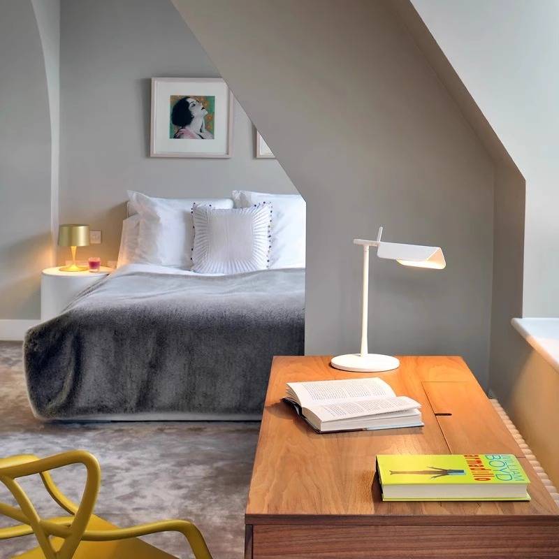 In a cozy bedroom with a slanted ceiling, a bed draped with a gray blanket and white pillows sits nearby. A wooden desk showcases an open book, the Simple Desktop Lamp, and a yellow book. A minimalist vibe is achieved with a yellow chair and nightstand featuring a modern lamp.
