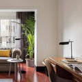 A modern living room features a cozy sofa with yellow pillows, adjacent to a wooden desk showcasing the Simple Desktop Lamp and books. A large potted plant adds greenery. Minimalist elements like floor-length curtains frame a window, offering city views on polished wood floors.