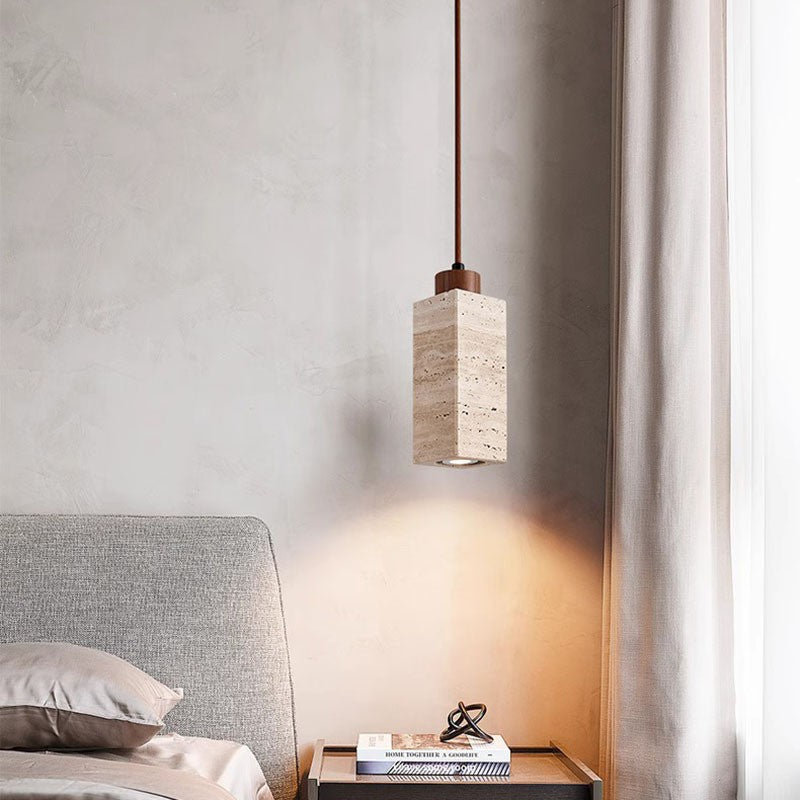 A bedroom with a textured beige wall includes a gray upholstered headboard. A square bedside table holds two books and a decorative metal object. Overhead, the Travertine Restaurant Chandelier with an adjustable wire and dimmable LED bulb softly illuminates the space.