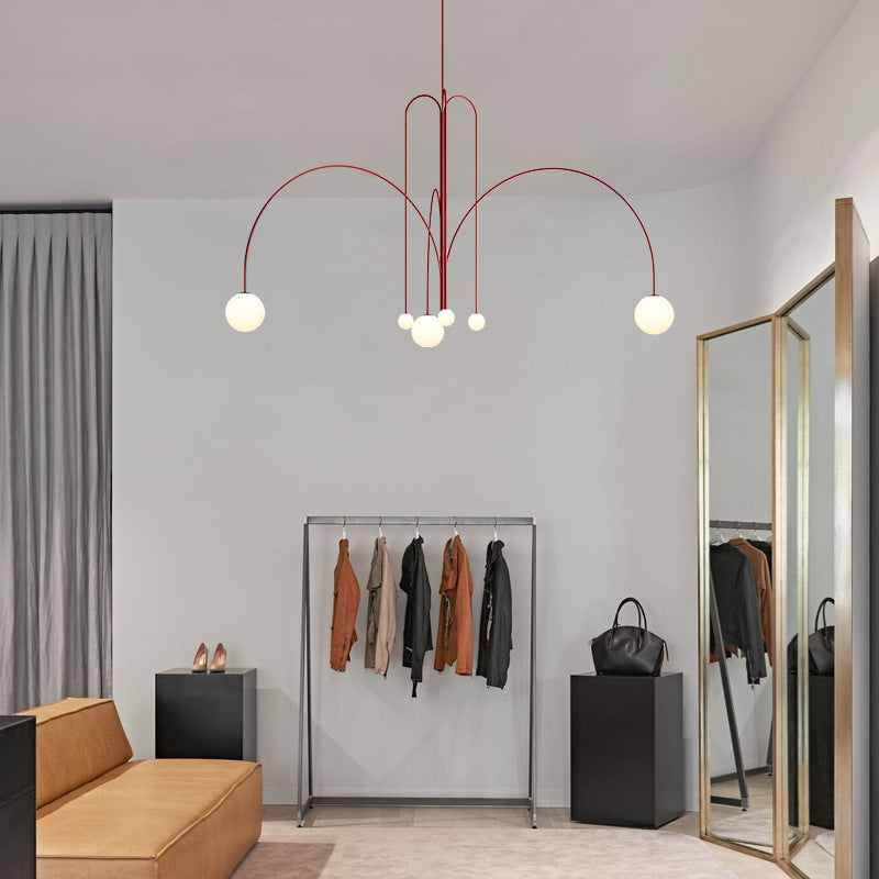 A modern room features the contemporary St Kilda Chandelier in red, a clothing rack with jackets and coats, a full-length mirror, and a brown sofa. A pair of shoes and a black handbag rest on nearby black pedestals against neutral walls and curtains.