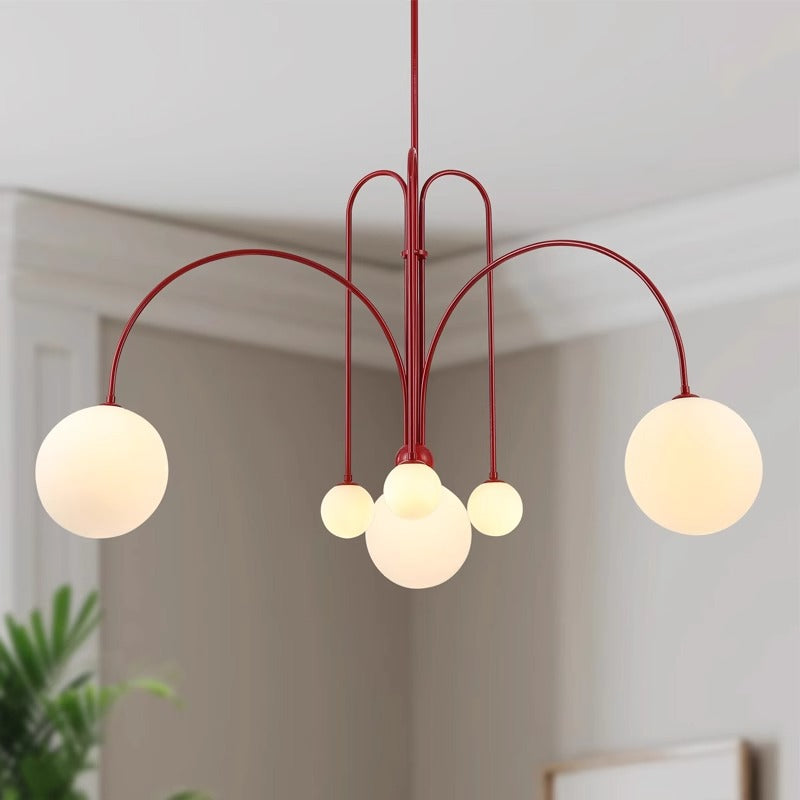 The St Kilda Chandelier features five illuminated, globe-shaped bulbs on red curved arms, suspended from the ceiling of a room with light walls and crown molding. A touch of green foliage decorates one corner.