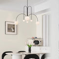 A modern kitchen includes a round white table with black chairs and a black vase filled with white tulips. Above the table, the St Kilda Chandelier with five round white lights is suspended. A microwave sits on the counter, and a framed photo adorns the wall.