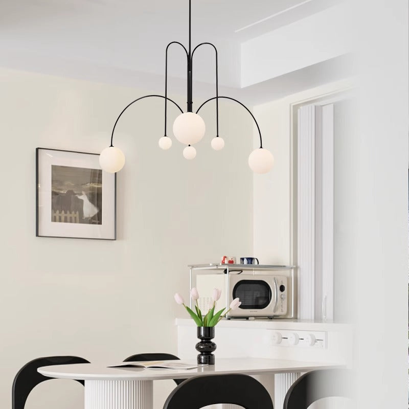 A modern kitchen includes a round white table with black chairs and a black vase filled with white tulips. Above the table, the St Kilda Chandelier with five round white lights is suspended. A microwave sits on the counter, and a framed photo adorns the wall.