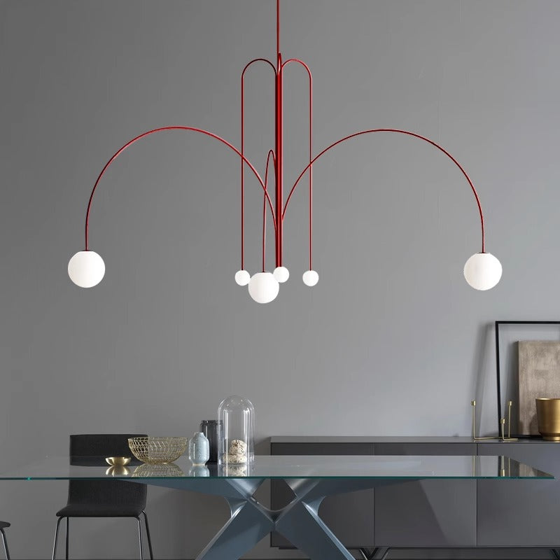 Modern dining room featuring a sleek glass table, decorative bowl, and a dome-covered item. Above hangs the St Kilda Chandelier with its abstract red design and white globes, contrasting against the gray wall. A framed picture is partially visible in the corner.