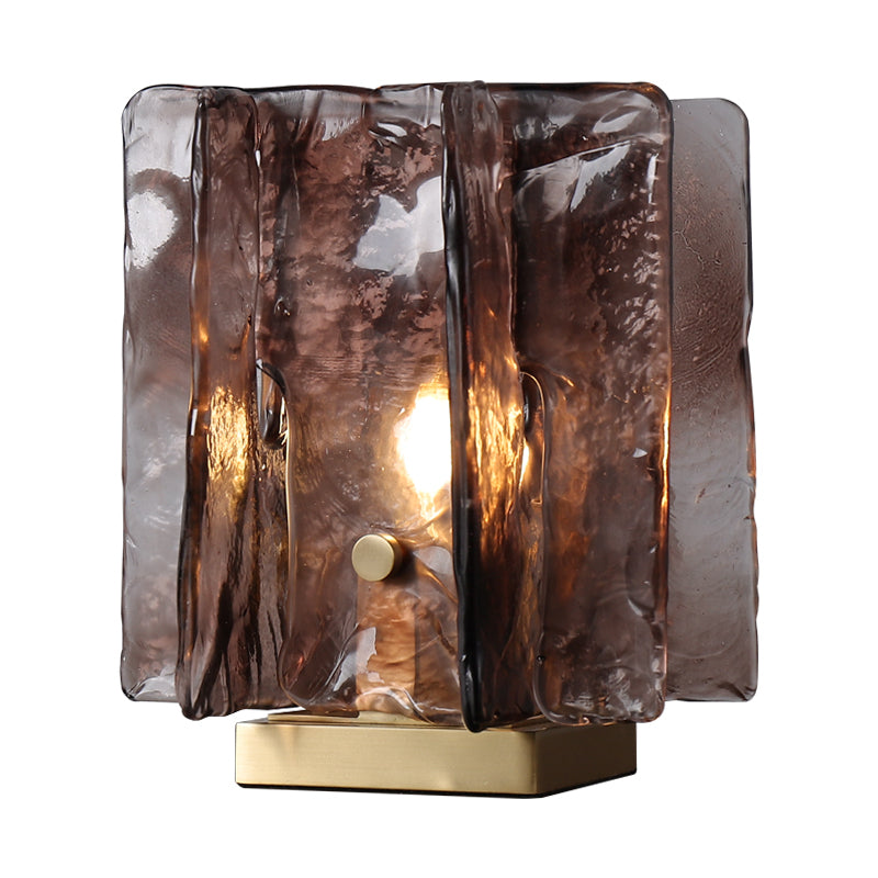The Vintage brass glass table lamp highlights a hand-blown amber shade in smoky brown, featuring vertical textured panels around a central bulb. Its supported by a rectangular gold base and emits warm, energy-efficient light through its translucent glass.