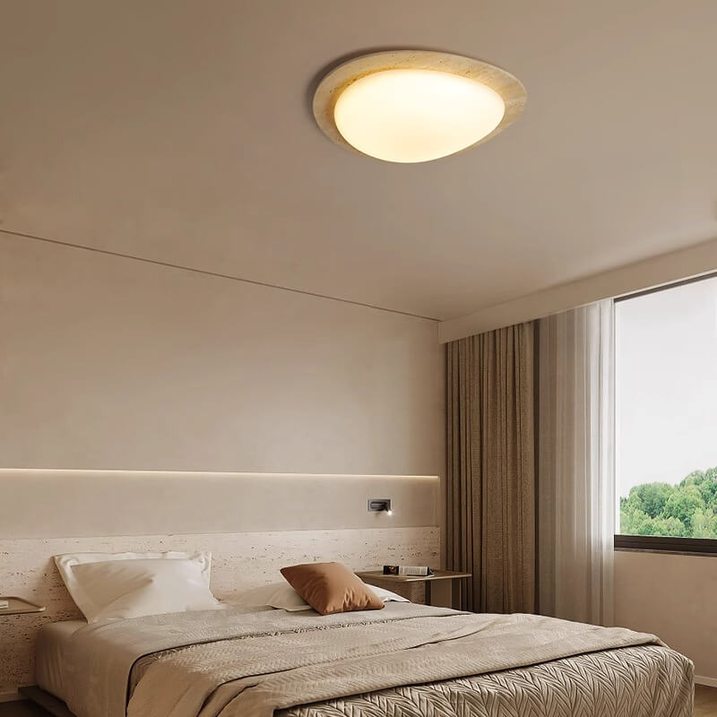 A modern bedroom with a large bed, neutral bedding, minimal decor, sheer curtains, a natural travertine bedside table, and the Wabi-Sabi Travertine LED Ceiling Light brightening the space with views of greenery outside.